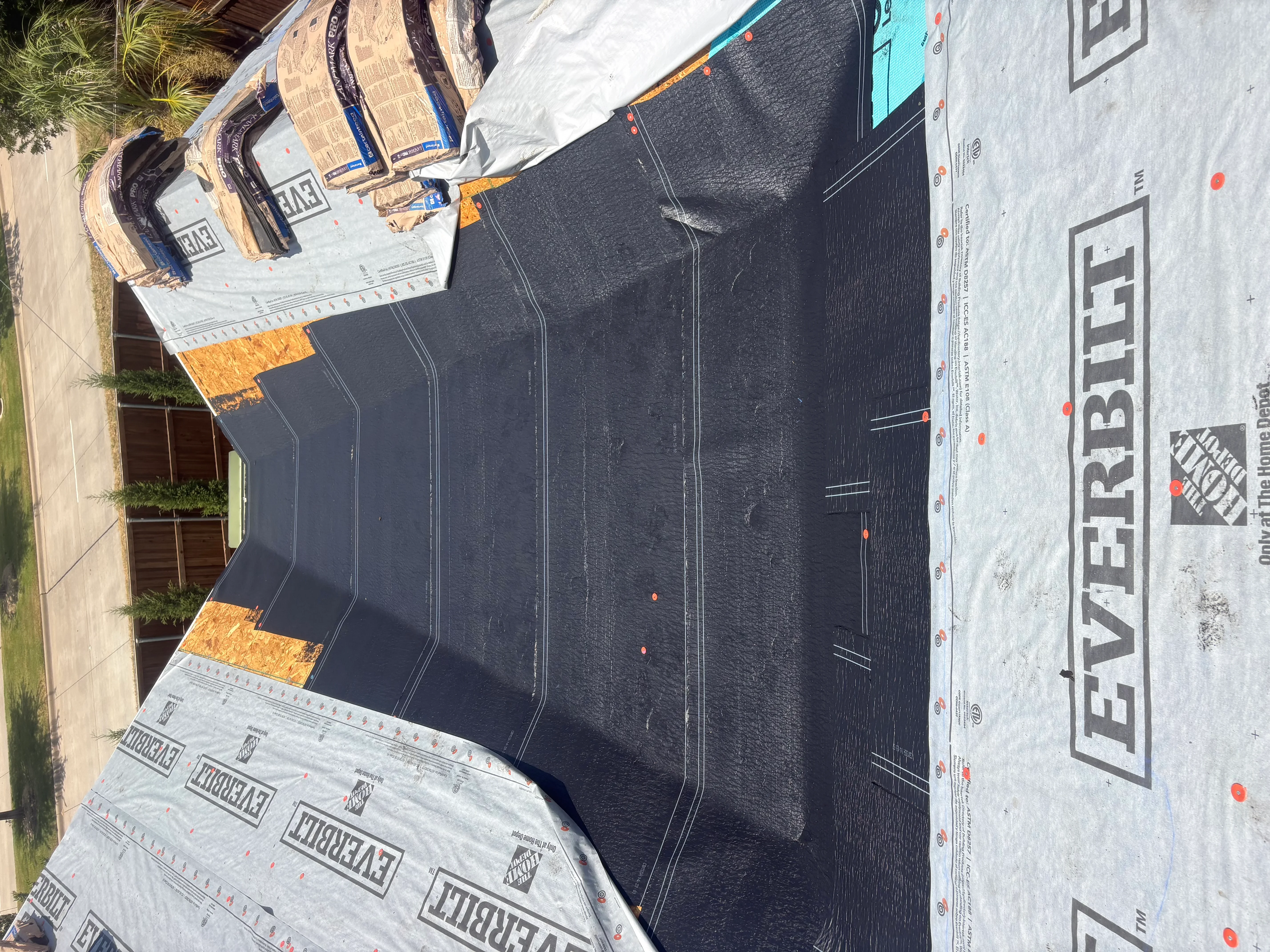 Black shingles with white underlayment, orange OSB sheathing, and metal drip edge during roof installation
